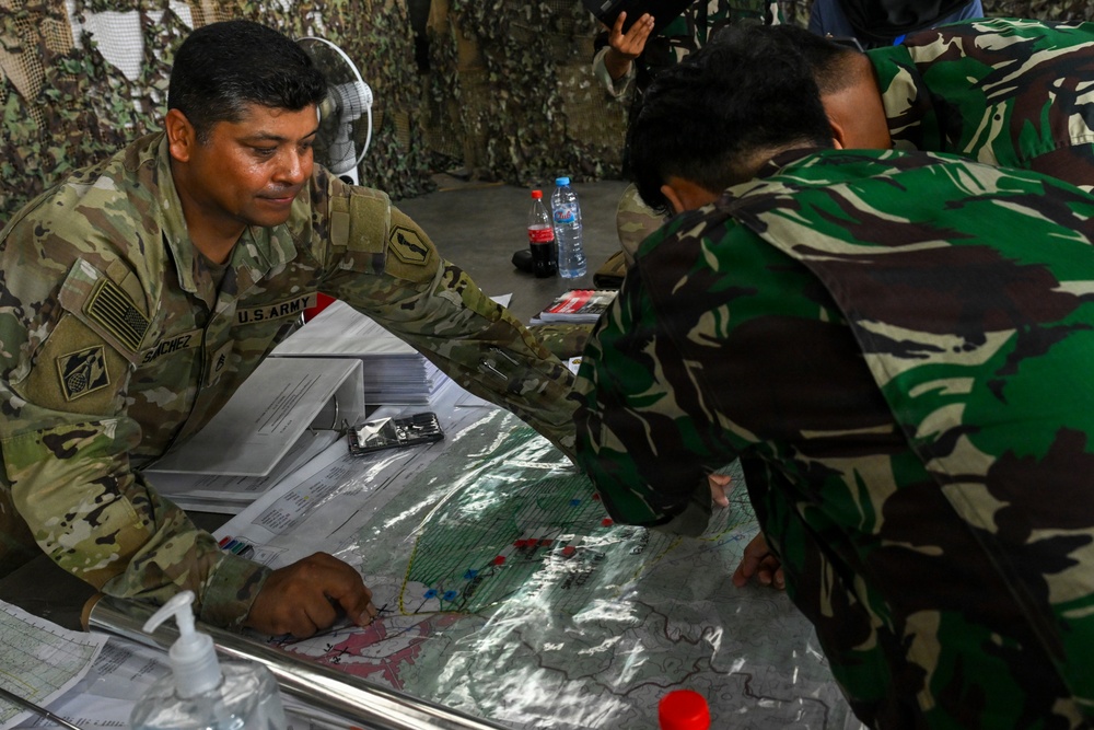 A U.S. Army soldier identified as 'Sanchez' is engaged in a joint military planning session with personnel wearing Indonesian Armed Forces (TNI) camouflage uniforms. They are gathered around a tactical map covered in plastic, pointing at various marked positions. The setting appears to be a field command post or operations tent with camouflage netting visible in the background.