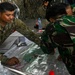 A U.S. Army soldier identified as 'Sanchez' is engaged in a joint military planning session with personnel wearing Indonesian Armed Forces (TNI) camouflage uniforms. They are gathered around a tactical map covered in plastic, pointing at various marked positions. The setting appears to be a field command post or operations tent with camouflage netting visible in the background.