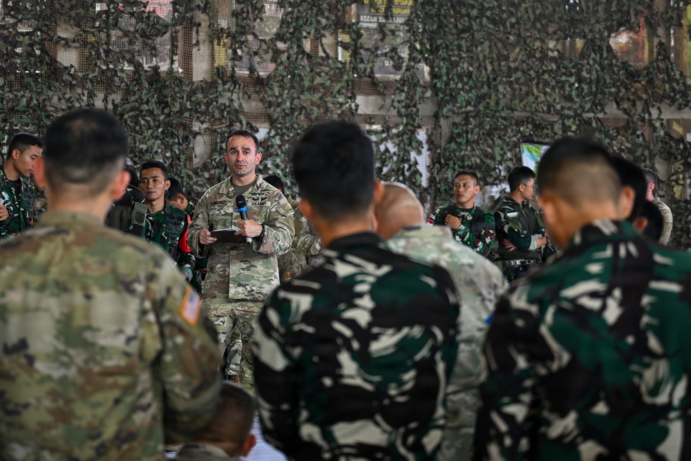 A U.S. Army soldier is addressing a mixed group of military personnel under a camouflage netting structure, holding a microphone and a notepad. The audience includes soldiers in U.S. Army OCP (Operational Camouflage Pattern) uniforms and others in a distinctive black-and-green camouflage pattern consistent with Indonesian Army (TNI-AD) uniforms. This appears to be a joint military briefing or training exercise between U.S. and Indonesian armed forces.
