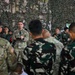 A U.S. Army soldier is addressing a mixed group of military personnel under a camouflage netting structure, holding a microphone and a notepad. The audience includes soldiers in U.S. Army OCP (Operational Camouflage Pattern) uniforms and others in a distinctive black-and-green camouflage pattern consistent with Indonesian Army (TNI-AD) uniforms. This appears to be a joint military briefing or training exercise between U.S. and Indonesian armed forces.
