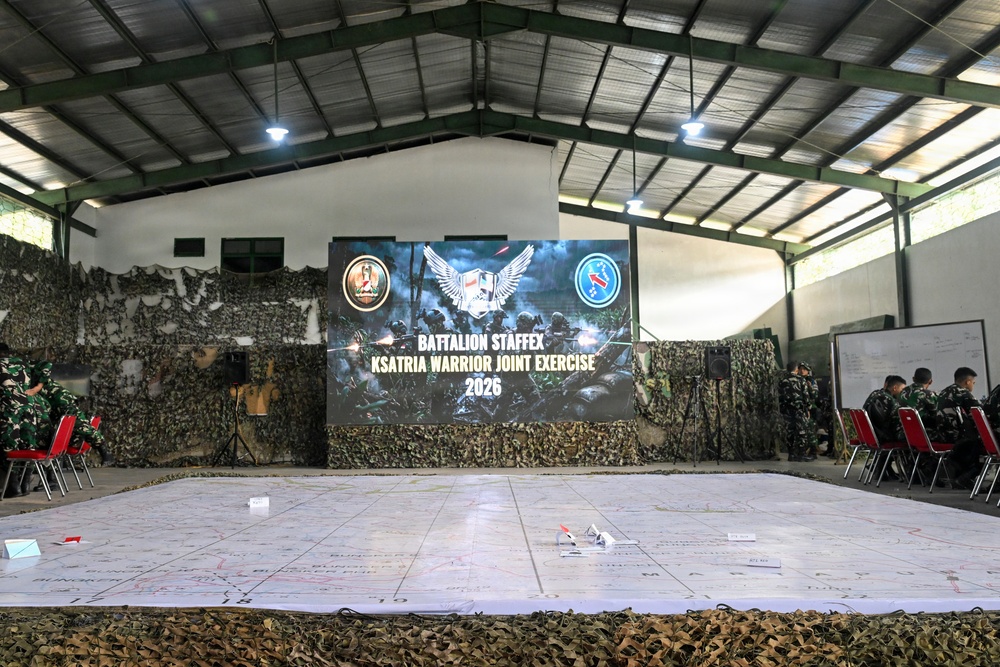 A military briefing or planning room is set up for the 'Battalion Staffex Ksatria Warrior Joint Exercise 2026,' featuring a large projected screen displaying the exercise title and military insignia. A large tactical map is spread across the floor in the foreground, and military personnel in camouflage uniforms are seated along the sides. The venue is decorated with camouflage netting and equipped with audio-visual equipment.