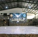 A military briefing or planning room is set up for the 'Battalion Staffex Ksatria Warrior Joint Exercise 2026,' featuring a large projected screen displaying the exercise title and military insignia. A large tactical map is spread across the floor in the foreground, and military personnel in camouflage uniforms are seated along the sides. The venue is decorated with camouflage netting and equipped with audio-visual equipment.