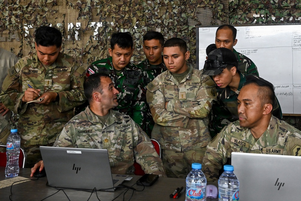 A group of U.S. Army soldiers and what appear to be Indonesian military personnel (TNI-AD) are gathered around laptops in a field operations center covered with camouflage netting. The session appears to be a joint military planning or training exercise, with personnel reviewing information on HP laptops and a whiteboard in the background. Name tapes visible include SANCHEZ, VARGAS, ACASIO, and VELASCO on U.S. Army uniforms.