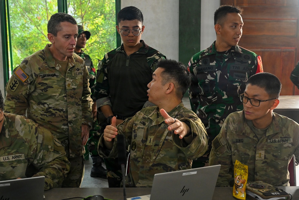 The image shows a joint military briefing or planning session involving U.S. Army personnel and Indonesian Armed Forces (TNI) soldiers. Several individuals are gathered around laptops, with one U.S. Army soldier gesturing and apparently explaining something to a senior officer. The setting appears to be an open-air or semi-outdoor facility, suggesting a multinational military exercise or coordination meeting.