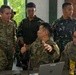 The image shows a joint military briefing or planning session involving U.S. Army personnel and Indonesian Armed Forces (TNI) soldiers. Several individuals are gathered around laptops, with one U.S. Army soldier gesturing and apparently explaining something to a senior officer. The setting appears to be an open-air or semi-outdoor facility, suggesting a multinational military exercise or coordination meeting.