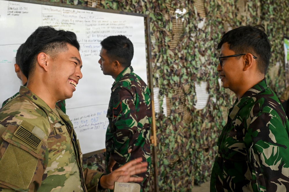 A U.S. Army soldier wearing OCP (Operational Camouflage Pattern) uniform with an American flag patch is engaged in a friendly conversation with military personnel wearing Indonesian Army (TNI-AD) camouflage uniforms. The setting appears to be a military briefing or planning area with a whiteboard and camouflage netting in the background. The interaction suggests a joint military exercise or training event between U.S. and Indonesian forces.