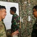 A U.S. Army soldier wearing OCP (Operational Camouflage Pattern) uniform with an American flag patch is engaged in a friendly conversation with military personnel wearing Indonesian Army (TNI-AD) camouflage uniforms. The setting appears to be a military briefing or planning area with a whiteboard and camouflage netting in the background. The interaction suggests a joint military exercise or training event between U.S. and Indonesian forces.