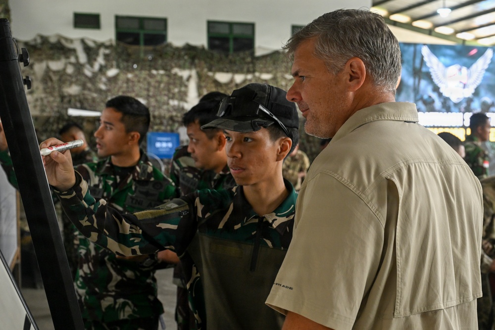 The image shows a group of military personnel in camouflage uniforms gathered around a whiteboard or easel in what appears to be an indoor briefing or training session. A civilian man in a khaki shirt stands alongside the uniformed individuals, observing as someone writes on the board. The setting includes camouflage netting in the background, suggesting a military operations or command environment.