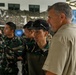 The image shows a group of military personnel in camouflage uniforms gathered around a whiteboard or easel in what appears to be an indoor briefing or training session. A civilian man in a khaki shirt stands alongside the uniformed individuals, observing as someone writes on the board. The setting includes camouflage netting in the background, suggesting a military operations or command environment.