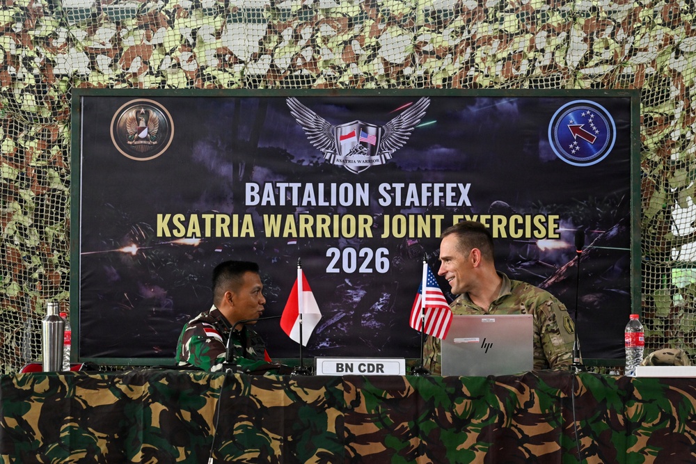 Two military officers — one in Indonesian Army camouflage uniform and one in U.S. Army camouflage uniform — are seated at a table during the Battalion STAFFEX Ksatria Warrior Joint Exercise 2026. The setting appears to be a military briefing or command post tent decorated with camouflage netting. A nameplate reading 'BN CDR' (Battalion Commander) is visible on the table between them.