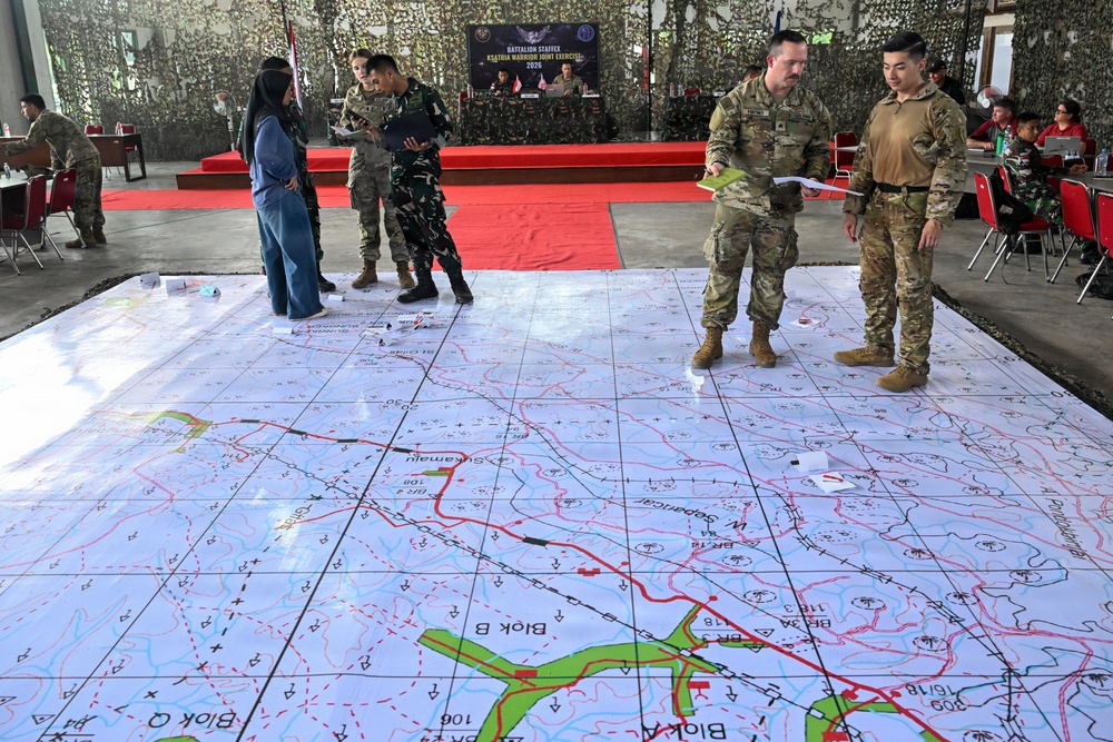 Military personnel from multiple nations are conducting a battalion staff exercise called 'Ksatria Warrior Joint Exercise 2026' in an open-air venue decorated with camouflage netting. Participants are gathered around a large floor-mounted tactical map displaying terrain features, roads, grid coordinates, and block designations. The exercise appears to be a joint planning or war-gaming session involving both U.S. and Indonesian military forces.