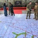 Military personnel from multiple nations are conducting a battalion staff exercise called 'Ksatria Warrior Joint Exercise 2026' in an open-air venue decorated with camouflage netting. Participants are gathered around a large floor-mounted tactical map displaying terrain features, roads, grid coordinates, and block designations. The exercise appears to be a joint planning or war-gaming session involving both U.S. and Indonesian military forces.