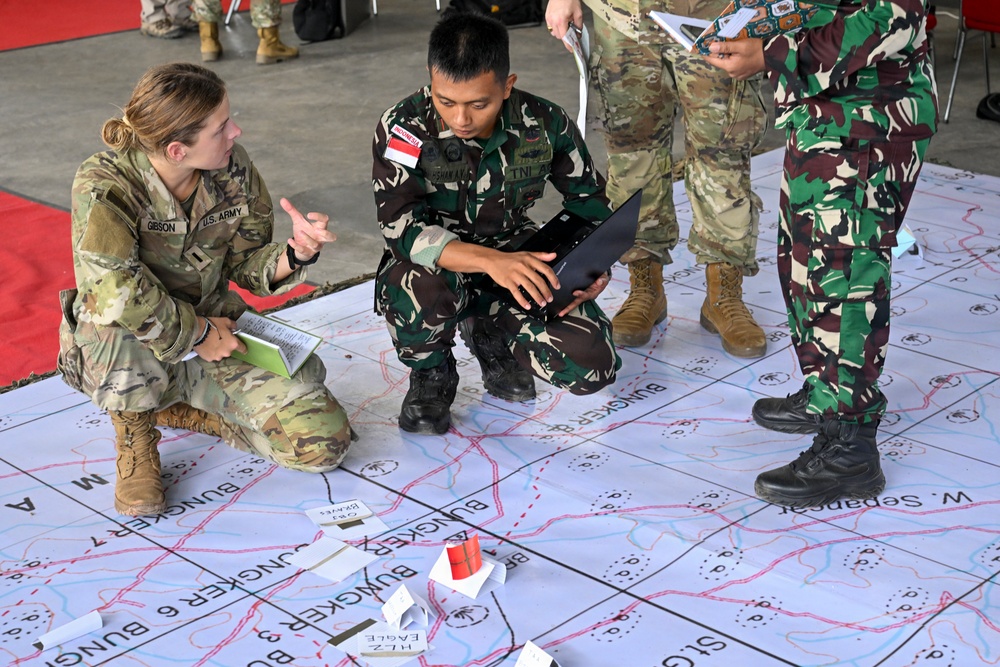 U.S. Army soldier (name tag: GIBSON) and Indonesian military personnel (TNI - Tentara Nasional Indonesia) are engaged in a joint military planning exercise. They are crouched over a large floor map marked with tactical designations including 'BUNGKER' positions, 'HLZ EAGLE,' and grid references. The exercise appears to be a combined arms or joint operational planning session between U.S. and Indonesian forces.