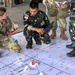 U.S. Army soldier (name tag: GIBSON) and Indonesian military personnel (TNI - Tentara Nasional Indonesia) are engaged in a joint military planning exercise. They are crouched over a large floor map marked with tactical designations including 'BUNGKER' positions, 'HLZ EAGLE,' and grid references. The exercise appears to be a combined arms or joint operational planning session between U.S. and Indonesian forces.