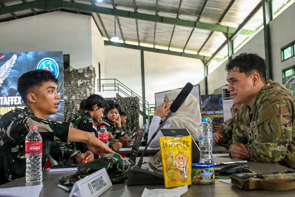 A military meeting or briefing session showing personnel from what appears to be two different armed forces seated across from each other at a table. One individual wears a U.S. Army multicam uniform while others wear Indonesian military (TNI) camouflage uniforms, suggesting a joint military exercise or staff exchange. A banner in the background partially reads 'STAFFEX' indicating a staff exercise event.