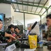 A military meeting or briefing session showing personnel from what appears to be two different armed forces seated across from each other at a table. One individual wears a U.S. Army multicam uniform while others wear Indonesian military (TNI) camouflage uniforms, suggesting a joint military exercise or staff exchange. A banner in the background partially reads 'STAFFEX' indicating a staff exercise event.