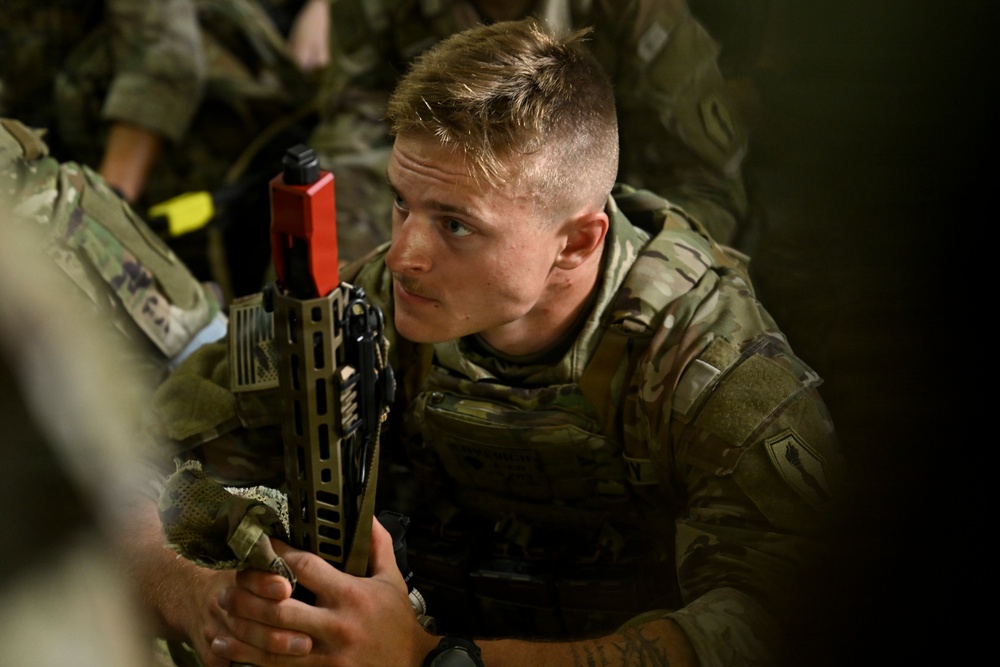 A young male soldier in MultiCam/OCP camouflage uniform is holding a rifle equipped with a red training bolt carrier group indicator, signifying a training or MILES gear configuration. He appears attentive, looking slightly off-camera, surrounded by other soldiers in similar uniforms. The setting appears to be an indoor or low-light training environment.