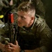 A young male soldier in MultiCam/OCP camouflage uniform is holding a rifle equipped with a red training bolt carrier group indicator, signifying a training or MILES gear configuration. He appears attentive, looking slightly off-camera, surrounded by other soldiers in similar uniforms. The setting appears to be an indoor or low-light training environment.