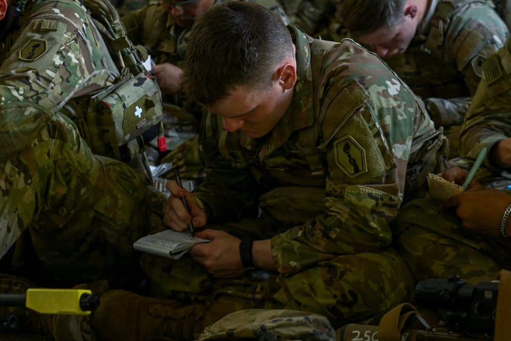 Multiple U.S. Army soldiers in Operational Camouflage Pattern (OCP) uniforms are seated closely together, with the central figure writing notes in a small notebook. The soldiers appear to be in a briefing or pre-mission preparation setting, possibly inside a military aircraft or staging area. Several soldiers are wearing tactical vests with visible medical and equipment pouches.