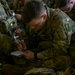 Multiple U.S. Army soldiers in Operational Camouflage Pattern (OCP) uniforms are seated closely together, with the central figure writing notes in a small notebook. The soldiers appear to be in a briefing or pre-mission preparation setting, possibly inside a military aircraft or staging area. Several soldiers are wearing tactical vests with visible medical and equipment pouches.