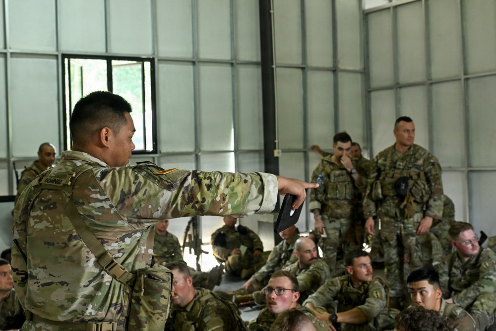 A U.S. Army soldier, identified by the name tape 'DOMINGO,' is conducting a briefing or instruction session for a large group of fellow soldiers seated on the floor inside a military hangar or open bay facility. The instructor is pointing toward the audience while holding what appears to be a notebook or tablet. The soldiers are wearing OCP (Operational Camouflage Pattern) uniforms, suggesting a contemporary U.S. Army training environment.