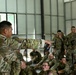 A U.S. Army soldier, identified by the name tape 'DOMINGO,' is conducting a briefing or instruction session for a large group of fellow soldiers seated on the floor inside a military hangar or open bay facility. The instructor is pointing toward the audience while holding what appears to be a notebook or tablet. The soldiers are wearing OCP (Operational Camouflage Pattern) uniforms, suggesting a contemporary U.S. Army training environment.