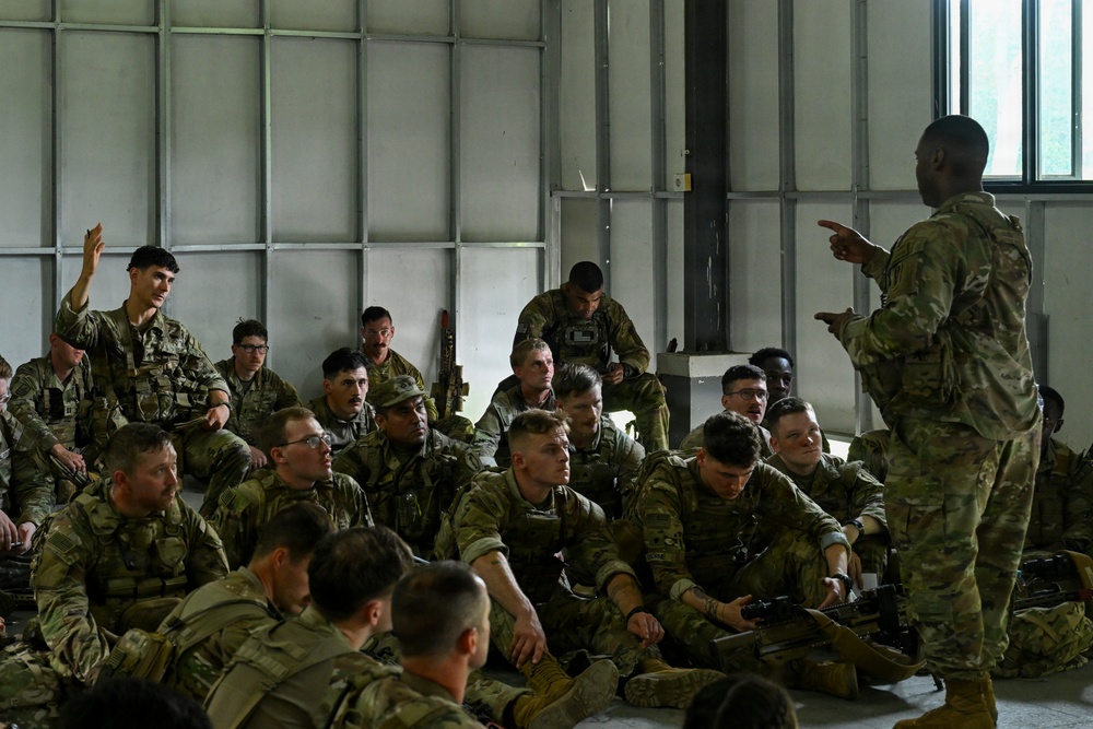 A military instructor or non-commissioned officer is conducting a briefing or after-action review with a large group of soldiers seated on the floor of what appears to be a hangar or warehouse facility. The soldiers are wearing OCP (Operational Camouflage Pattern) uniforms and are attentively listening, with at least one soldier raising their hand to ask a question. The setting suggests a training environment, with some soldiers holding rifles.