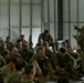 A military instructor or non-commissioned officer is conducting a briefing or after-action review with a large group of soldiers seated on the floor of what appears to be a hangar or warehouse facility. The soldiers are wearing OCP (Operational Camouflage Pattern) uniforms and are attentively listening, with at least one soldier raising their hand to ask a question. The setting suggests a training environment, with some soldiers holding rifles.