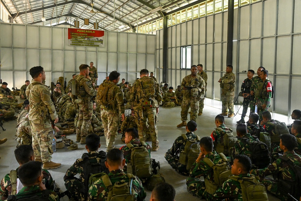 A military briefing or training session is taking place inside a large hangar or warehouse facility. Soldiers in two distinct camouflage uniforms — multicam/OCP pattern and a darker woodland pattern — are gathered together, with some standing and addressing the group while others sit on the floor. A banner on the wall reads 'PANCASILA,' indicating an Indonesian military connection.
