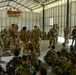 A military briefing or training session is taking place inside a large hangar or warehouse facility. Soldiers in two distinct camouflage uniforms — multicam/OCP pattern and a darker woodland pattern — are gathered together, with some standing and addressing the group while others sit on the floor. A banner on the wall reads 'PANCASILA,' indicating an Indonesian military connection.