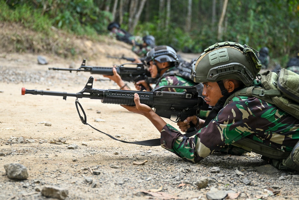 Multiple soldiers in camouflage uniforms are lying in a prone firing position on a dirt/gravel surface, aiming rifles downrange. The soldiers are wearing tactical helmets and carrying backpacks, suggesting a military training exercise. The setting appears to be an outdoor range or field training area with dense vegetation in the background.
