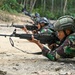 Multiple soldiers in camouflage uniforms are lying in a prone firing position on a dirt/gravel surface, aiming rifles downrange. The soldiers are wearing tactical helmets and carrying backpacks, suggesting a military training exercise. The setting appears to be an outdoor range or field training area with dense vegetation in the background.