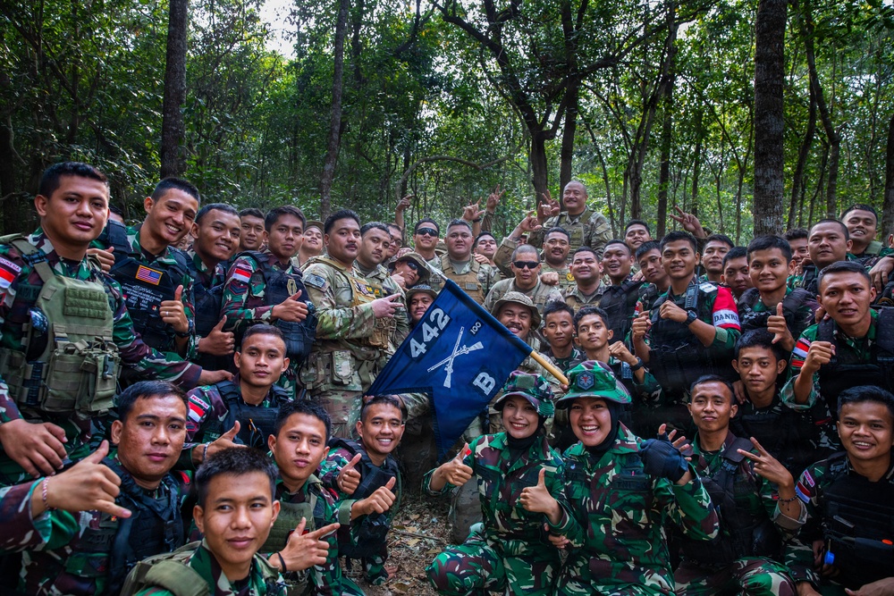 A large group of military personnel pose together in a forested area, displaying camaraderie and positive gestures such as thumbs up and peace signs. The group includes soldiers in Indonesian camouflage uniforms (TNI - Tentara Nasional Indonesia) alongside personnel in US military multicam uniforms. A blue pennant flag bearing the number '442' and crossed rifles insignia is prominently displayed in the center of the group.