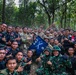 A large group of military personnel pose together in a forested area, displaying camaraderie and positive gestures such as thumbs up and peace signs. The group includes soldiers in Indonesian camouflage uniforms (TNI - Tentara Nasional Indonesia) alongside personnel in US military multicam uniforms. A blue pennant flag bearing the number '442' and crossed rifles insignia is prominently displayed in the center of the group.