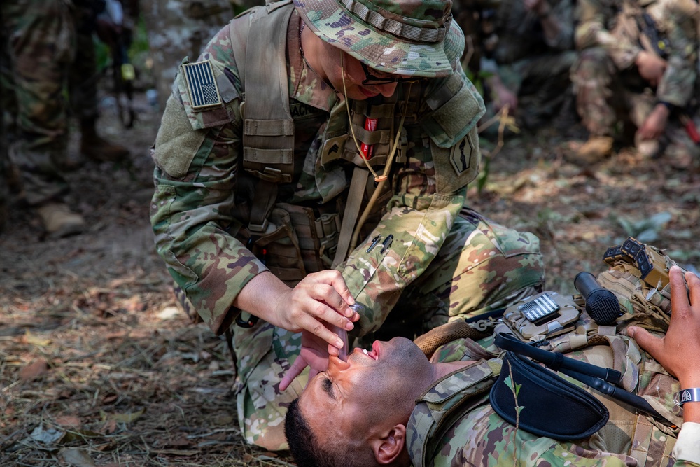 A U.S. Army soldier in OCP (Operational Camouflage Pattern) uniform is performing tactical combat casualty care (TCCC) on a fellow soldier lying on the ground. The kneeling soldier appears to be conducting an airway assessment or nasopharyngeal airway (NPA) insertion on the supine casualty. This appears to be a field medical training exercise based on the controlled setting and presence of observers.