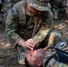 A U.S. Army soldier in OCP (Operational Camouflage Pattern) uniform is performing tactical combat casualty care (TCCC) on a fellow soldier lying on the ground. The kneeling soldier appears to be conducting an airway assessment or nasopharyngeal airway (NPA) insertion on the supine casualty. This appears to be a field medical training exercise based on the controlled setting and presence of observers.