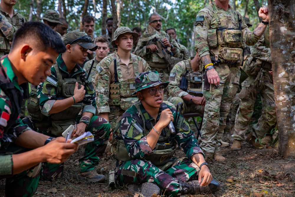 A multinational military training exercise is taking place in a forested area, with soldiers from at least two different nations visible. An Indonesian Armed Forces (TNI) soldier wearing a bucket hat and glasses is seated on the ground speaking into a microphone, while US Army soldiers in MultiCam uniforms stand and crouch nearby. The scene appears to be a briefing or after-action review conducted outdoors.