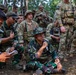 A multinational military training exercise is taking place in a forested area, with soldiers from at least two different nations visible. An Indonesian Armed Forces (TNI) soldier wearing a bucket hat and glasses is seated on the ground speaking into a microphone, while US Army soldiers in MultiCam uniforms stand and crouch nearby. The scene appears to be a briefing or after-action review conducted outdoors.