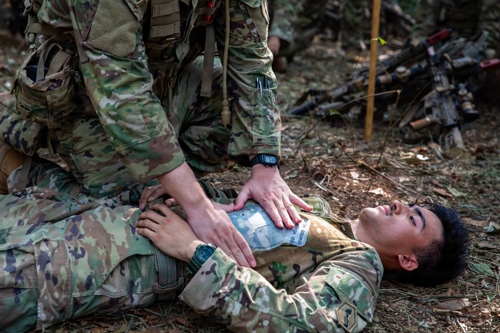 Military personnel are conducting a tactical combat casualty care (TCCC) training exercise in a wooded outdoor environment. One soldier is lying on the ground in a simulated casualty role while another soldier kneels over them applying what appears to be a chest seal or vented chest dressing to the torso area. The scene includes military equipment and weapons visible in the background, consistent with a field training exercise.
