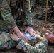 Military personnel are conducting a tactical combat casualty care (TCCC) training exercise in a wooded outdoor environment. One soldier is lying on the ground in a simulated casualty role while another soldier kneels over them applying what appears to be a chest seal or vented chest dressing to the torso area. The scene includes military equipment and weapons visible in the background, consistent with a field training exercise.