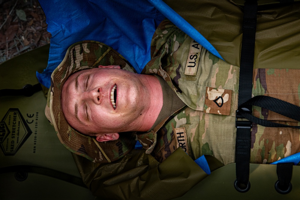 A U.S. Army soldier in OCP (Operational Camouflage Pattern) uniform is lying on a military-issue folding stretcher, appearing to simulate a casualty during a medical training exercise. The soldier's name tape reads 'URTH' (partially visible) and the 'U.S. AR[MY]' tape is visible on the chest. The stretcher is identifiable as a NATO-standard folding field stretcher with CE certification markings.