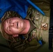 A U.S. Army soldier in OCP (Operational Camouflage Pattern) uniform is lying on a military-issue folding stretcher, appearing to simulate a casualty during a medical training exercise. The soldier's name tape reads 'URTH' (partially visible) and the 'U.S. AR[MY]' tape is visible on the chest. The stretcher is identifiable as a NATO-standard folding field stretcher with CE certification markings.