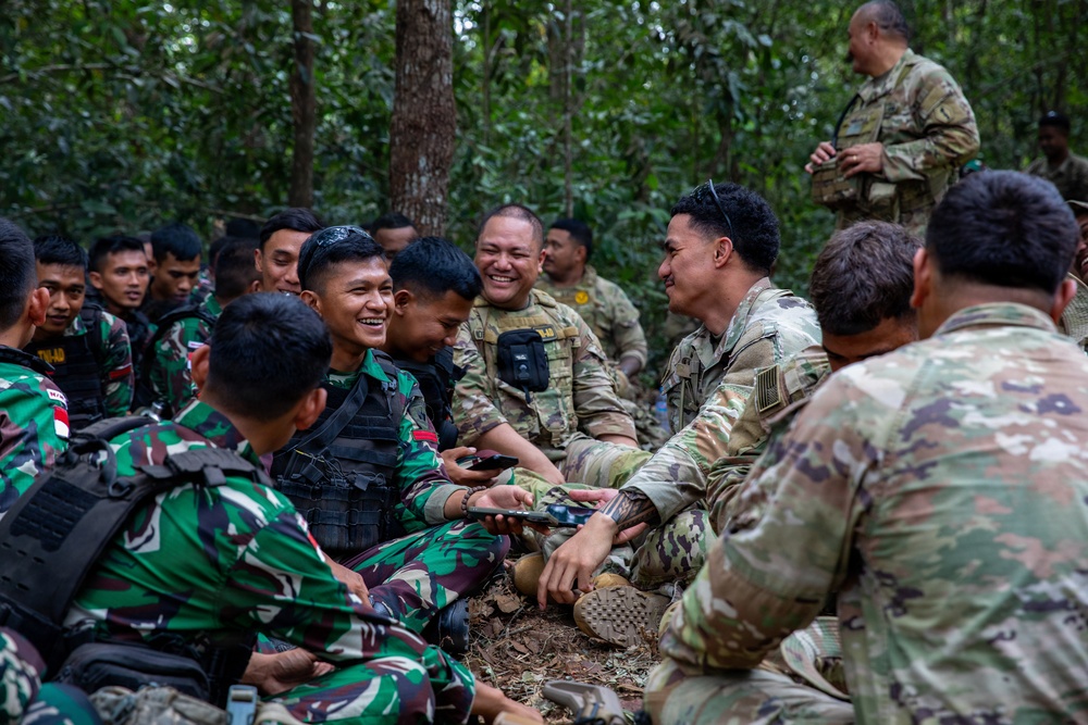 A group of military personnel from two different forces are gathered in a forested area, engaging in friendly interaction. Some soldiers are wearing Indonesian Army (TNI-AD) camouflage uniforms with Indonesian flag patches, while others wear US Army MultiCam/OCP uniforms. The atmosphere appears relaxed and positive, suggesting a joint training exercise or military exchange program.
