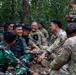 A group of military personnel from two different forces are gathered in a forested area, engaging in friendly interaction. Some soldiers are wearing Indonesian Army (TNI-AD) camouflage uniforms with Indonesian flag patches, while others wear US Army MultiCam/OCP uniforms. The atmosphere appears relaxed and positive, suggesting a joint training exercise or military exchange program.