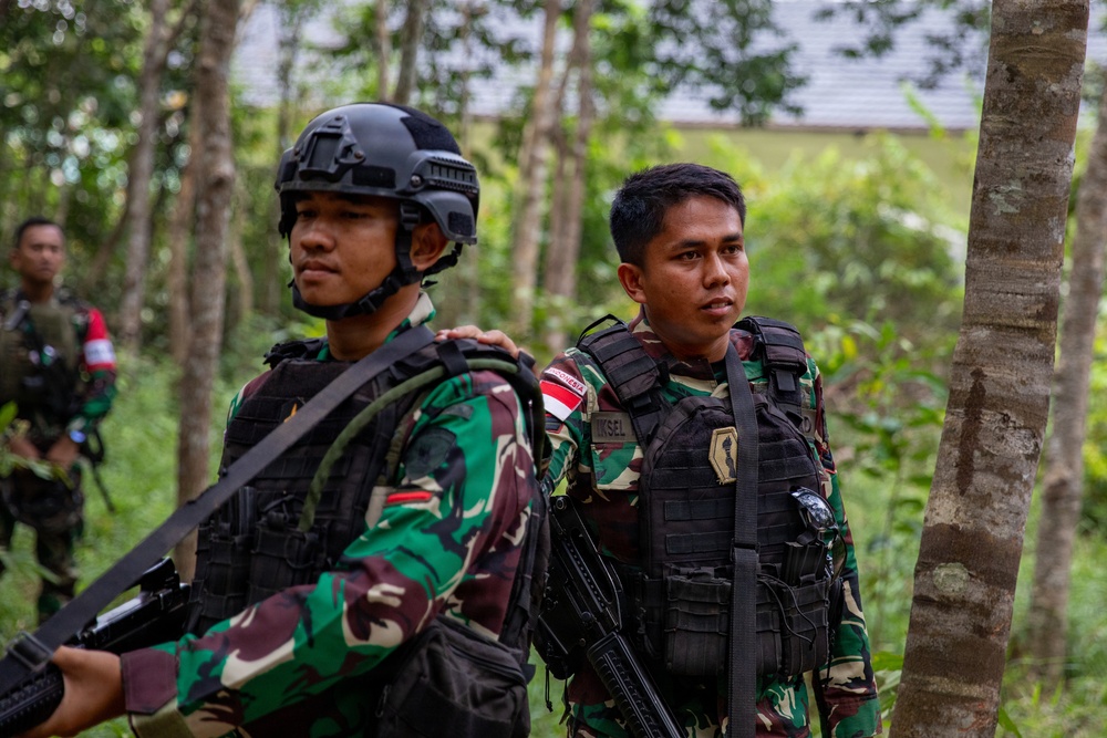 The image shows two Indonesian military personnel (TNI - Tentara Nasional Indonesia) in a forested area wearing camouflage uniforms and tactical gear. Both soldiers are equipped with body armor, weapons, and tactical accessories. A third soldier is partially visible in the background.