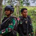 The image shows two Indonesian military personnel (TNI - Tentara Nasional Indonesia) in a forested area wearing camouflage uniforms and tactical gear. Both soldiers are equipped with body armor, weapons, and tactical accessories. A third soldier is partially visible in the background.