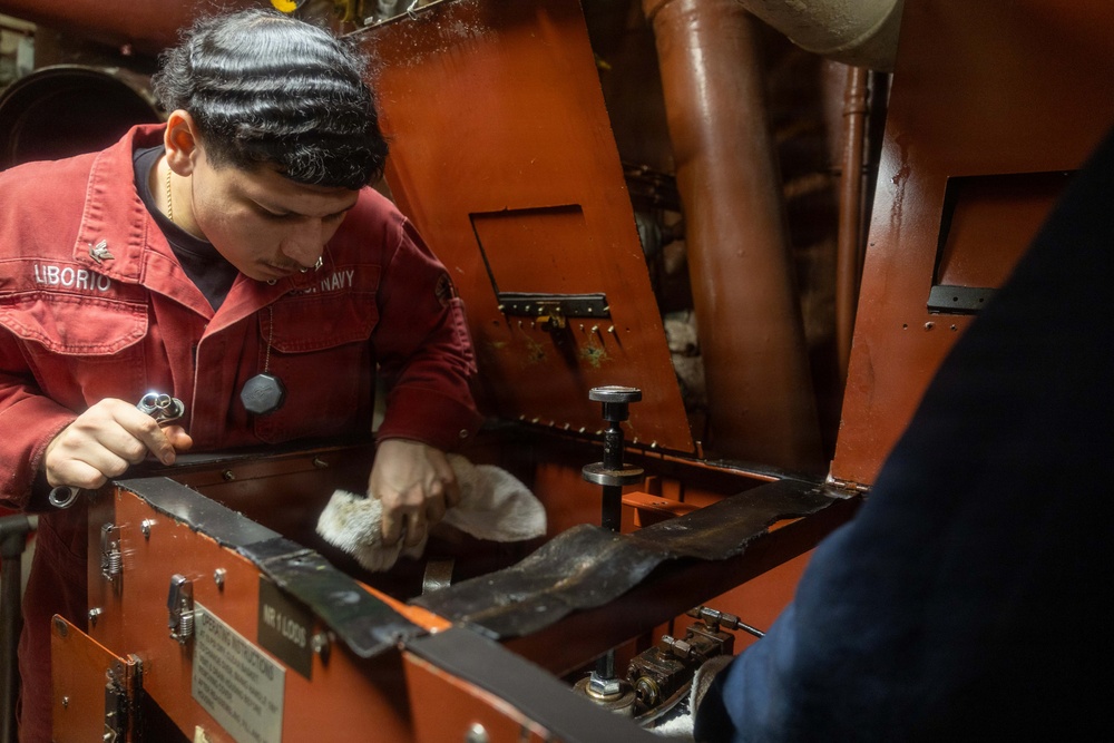 A U.S. Navy sailor named Liborio, wearing a red coverall uniform, is performing maintenance work on a piece of orange-painted machinery in what appears to be a ship's engineering or mechanical space. The sailor is using a small flashlight and a cleaning cloth to inspect or clean internal components of the equipment. A second person, partially visible on the right, appears to be assisting or observing.