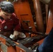A U.S. Navy sailor named Liborio, wearing a red coverall uniform, is performing maintenance work on a piece of orange-painted machinery in what appears to be a ship's engineering or mechanical space. The sailor is using a small flashlight and a cleaning cloth to inspect or clean internal components of the equipment. A second person, partially visible on the right, appears to be assisting or observing.