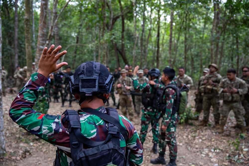 A group of military personnel are gathered in a forested area, appearing to be conducting a joint training exercise or briefing. Two soldiers in green camouflage uniforms consistent with Indonesian Army (TNI) are in the foreground, one with a raised hand, facing a mixed group that includes soldiers in both green camouflage and tan/multicam uniforms. The setting suggests a multinational military exercise in a tropical forest environment.