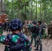 A group of military personnel are gathered in a forested area, appearing to be conducting a joint training exercise or briefing. Two soldiers in green camouflage uniforms consistent with Indonesian Army (TNI) are in the foreground, one with a raised hand, facing a mixed group that includes soldiers in both green camouflage and tan/multicam uniforms. The setting suggests a multinational military exercise in a tropical forest environment.